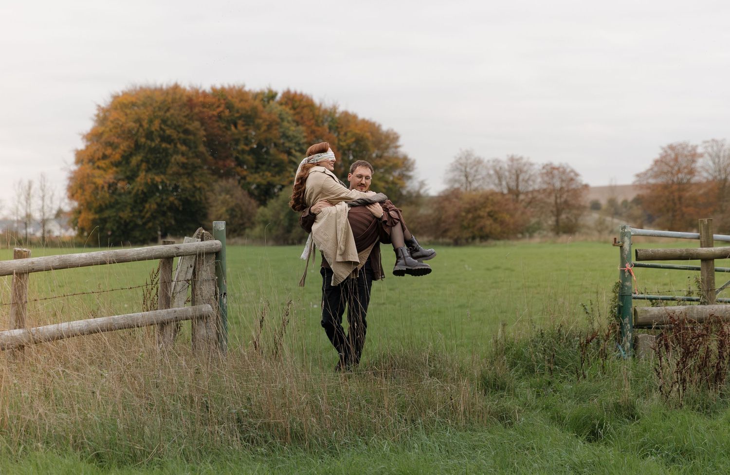 A panoramic view of people walking through a green autumn field with wooden fences and trees in the background.