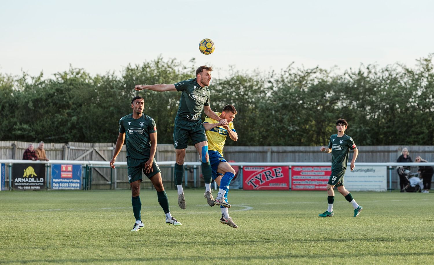Football players compete for a header during a match, with green-kitted players jumping near the sideline.