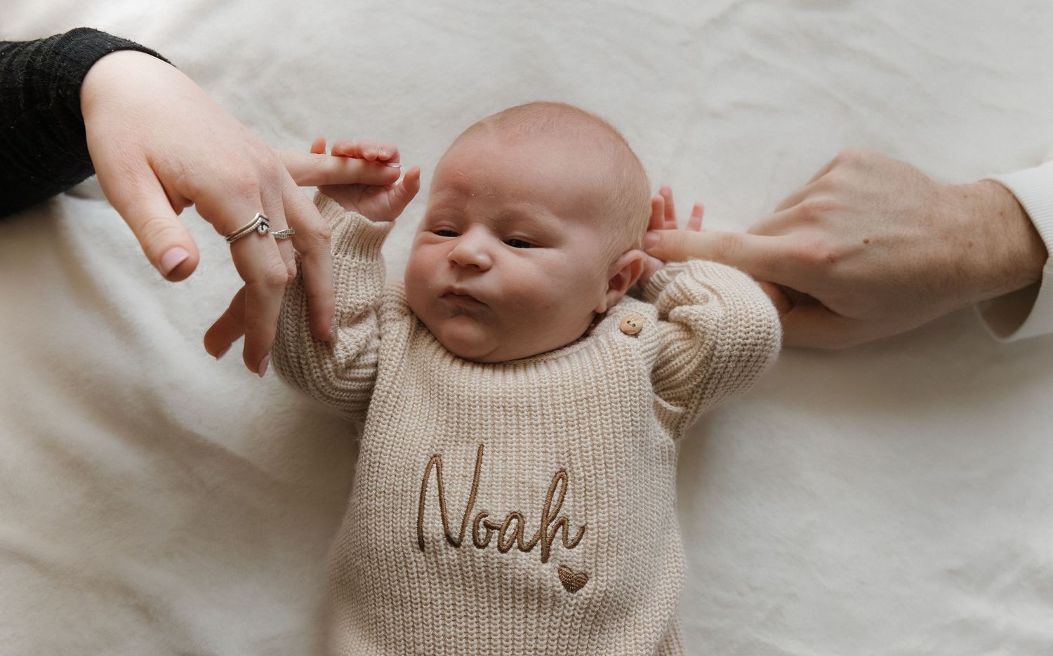 A baby wearing a cream knit sweater with 'Noah' embroidered on it lies on white fabric while hands reach to touch.