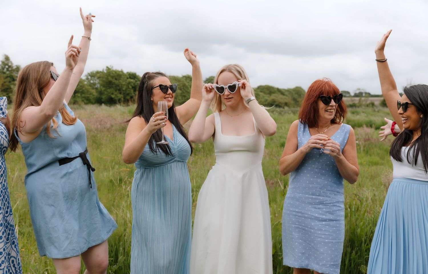 Group in blue dresses raise their hands in celebration against cloudy sky.