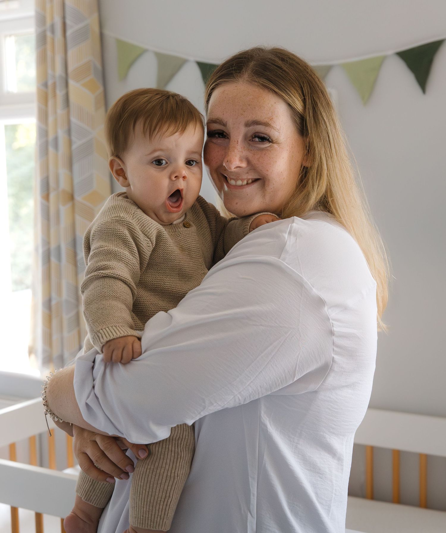 A parent in a white shirt holds and hugs a small child wearing a beige sweater in a bright room with bunting decor.