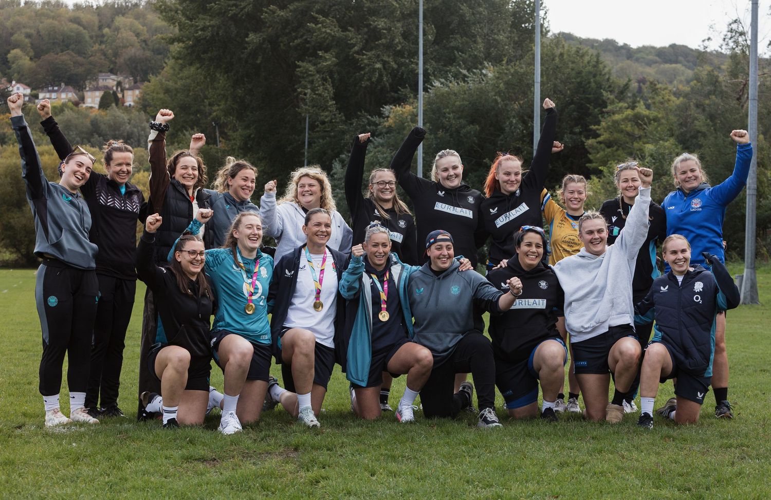 A group of bath rugby ladies athletes celebrates together on a sports field with raised arms and excited expressions.