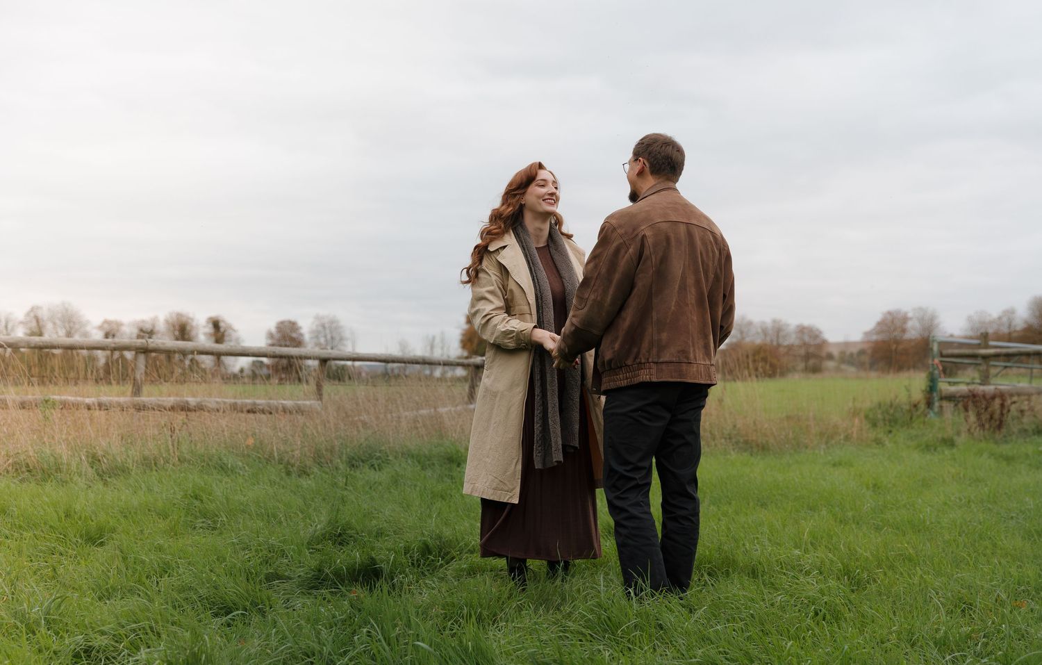 A couple walks together through a scenic meadow on an autumn afternoon with bare trees in the distance.