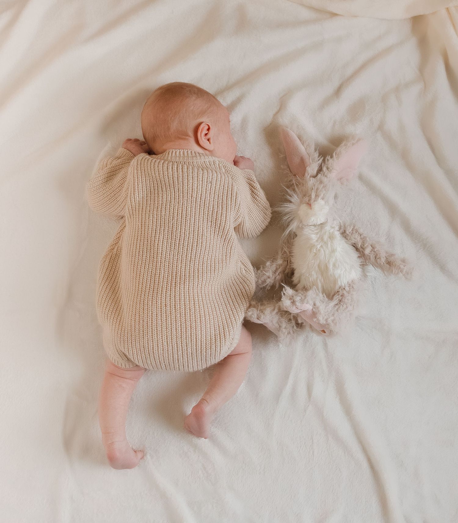 A sleeping baby in a cream knit outfit lies peacefully on soft white bedding.
