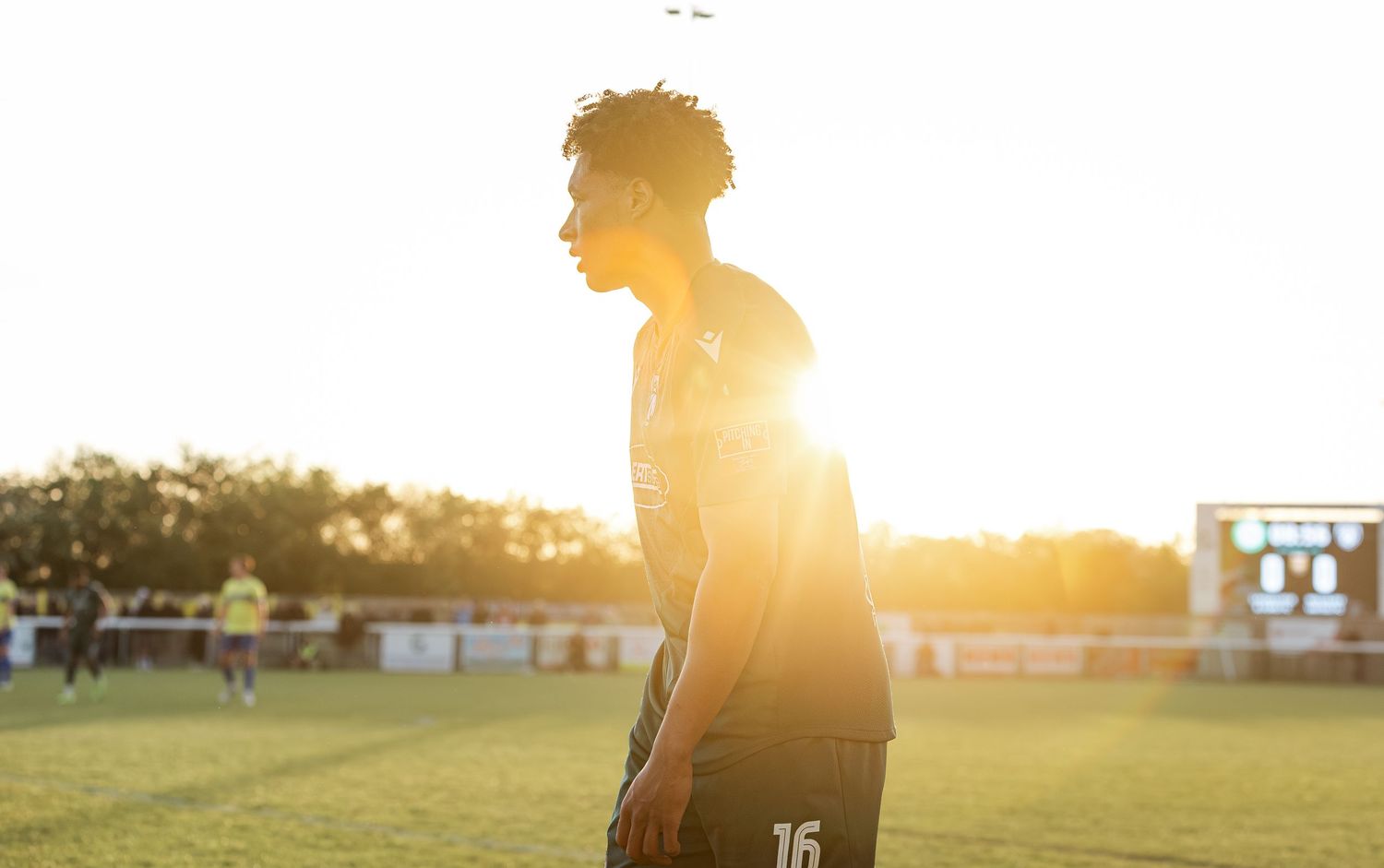Soccer player wearing number 16 jersey stands on field bathed in golden sunset light during outdoor match.