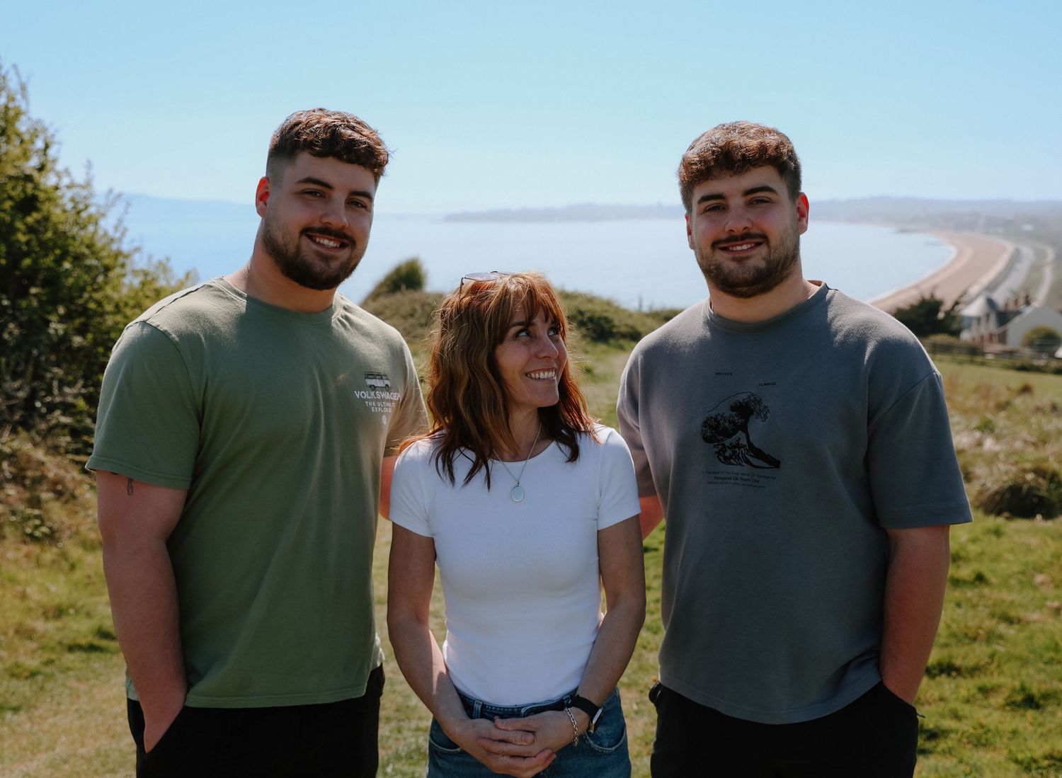 Three people posing together on grassy hillside on sunny day.