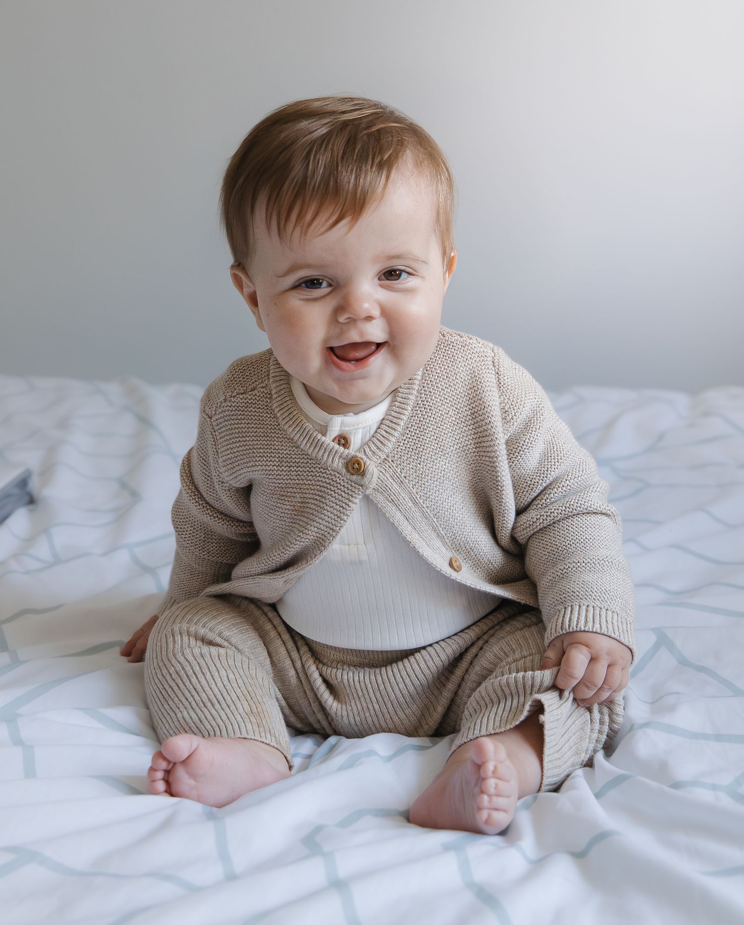 Baby wearing a cozy beige knit sweater and pants set sits on white bedding while smiling and playing.