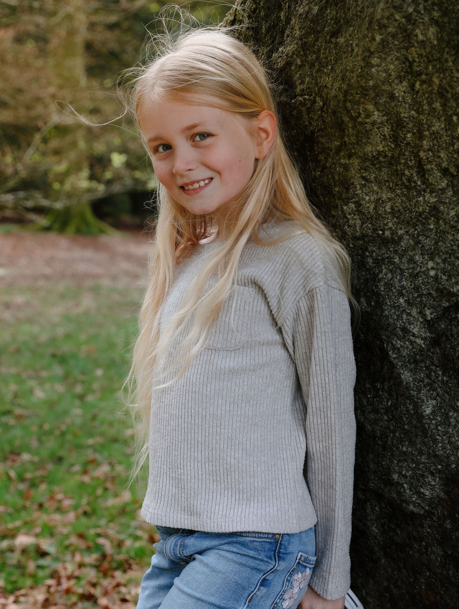 Young person in gray sweater leaning against tree trunk in autumn setting.