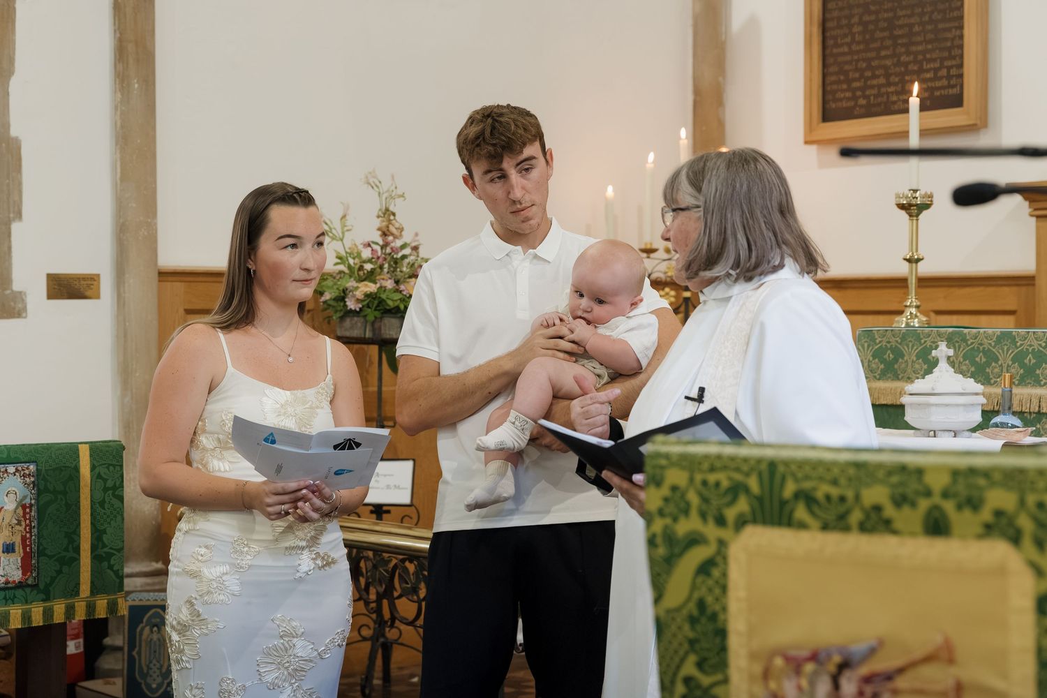 Young couple holding baby during christening ceremony with female officiant in church.
