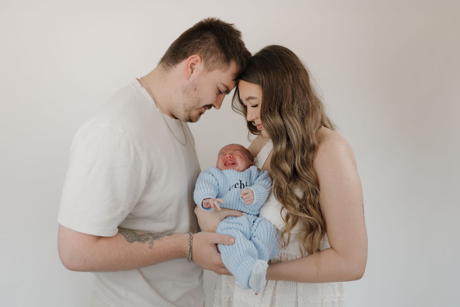 New parents tenderly hold their sleeping newborn baby wrapped in a blue swaddle against a white background.