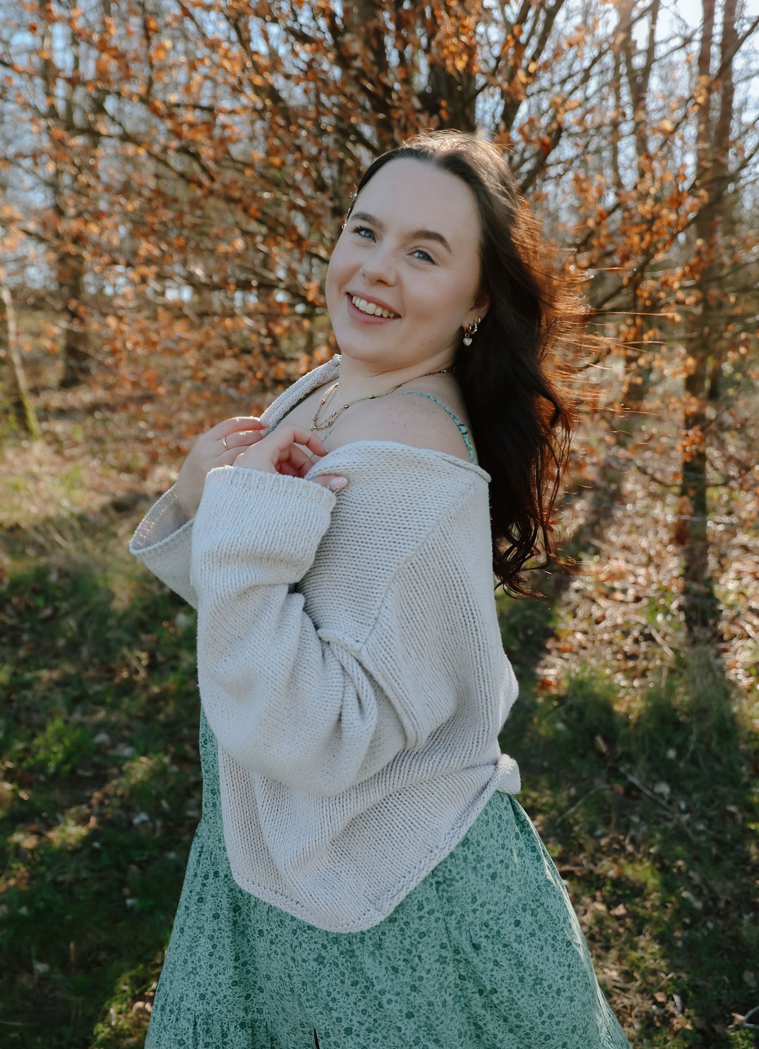 A person in a beige sweater and mint green skirt poses outdoors against autumn foliage.