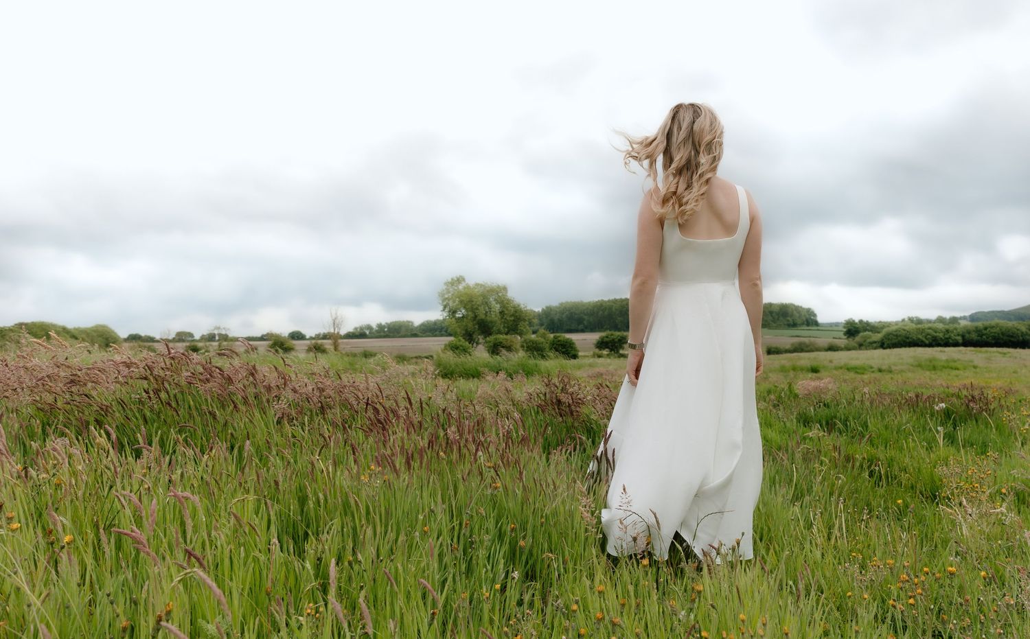 Woman in white dress stands in tall grass field under cloudy sky.