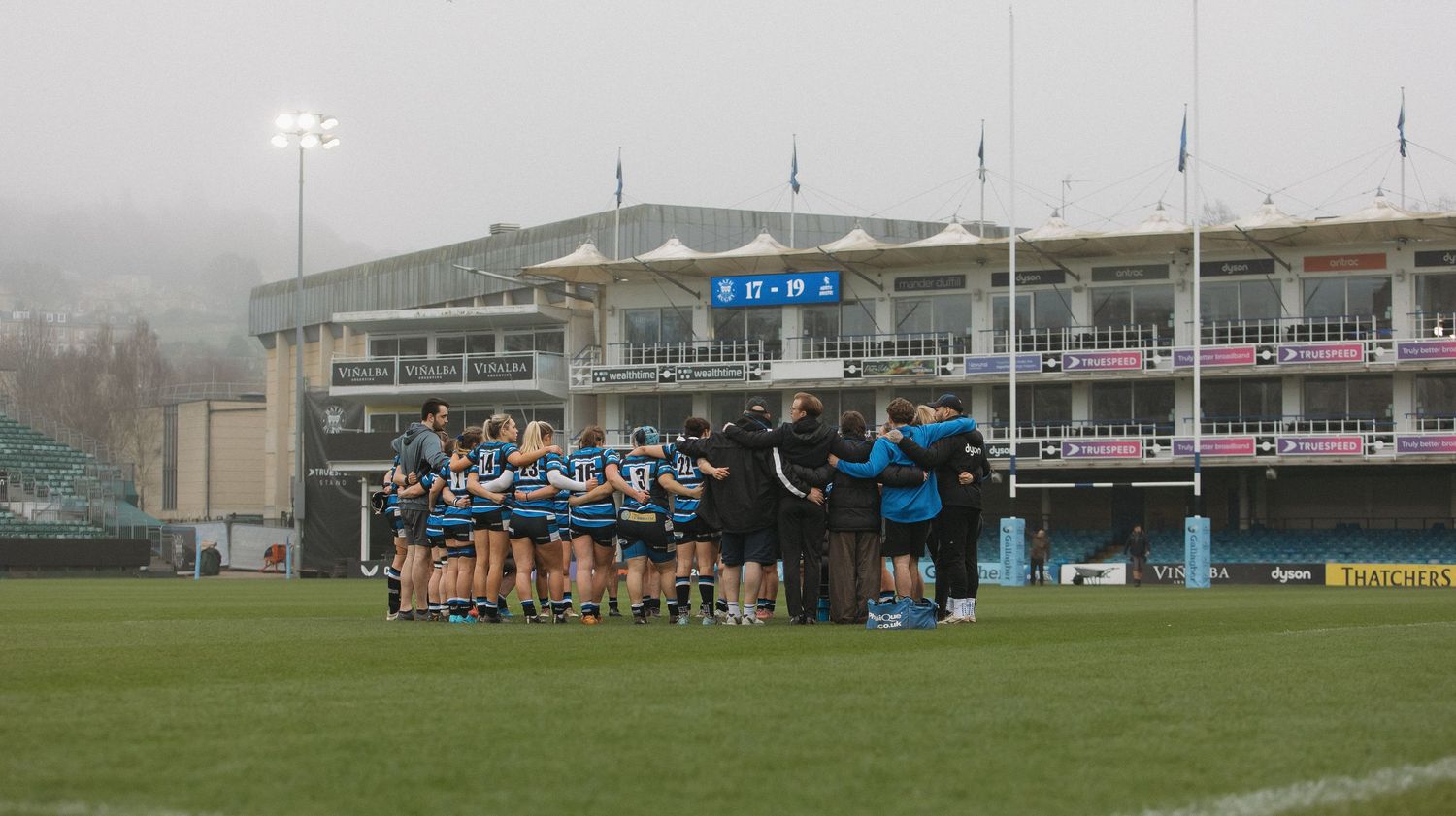 Rugby players gather on a misty training field with floodlights illuminating the outdoor sports facility.