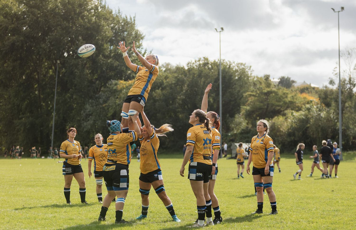 Bath rugby players in yellow jerseys compete for the ball during a practise line-out play on a grass field.