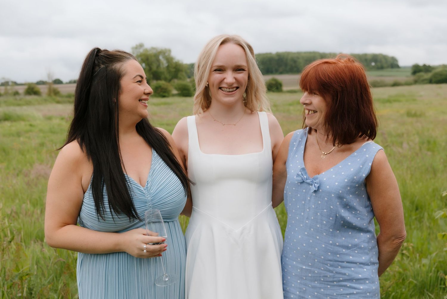 Group wearing coordinated light blue dresses laugh together in a sunny meadow.