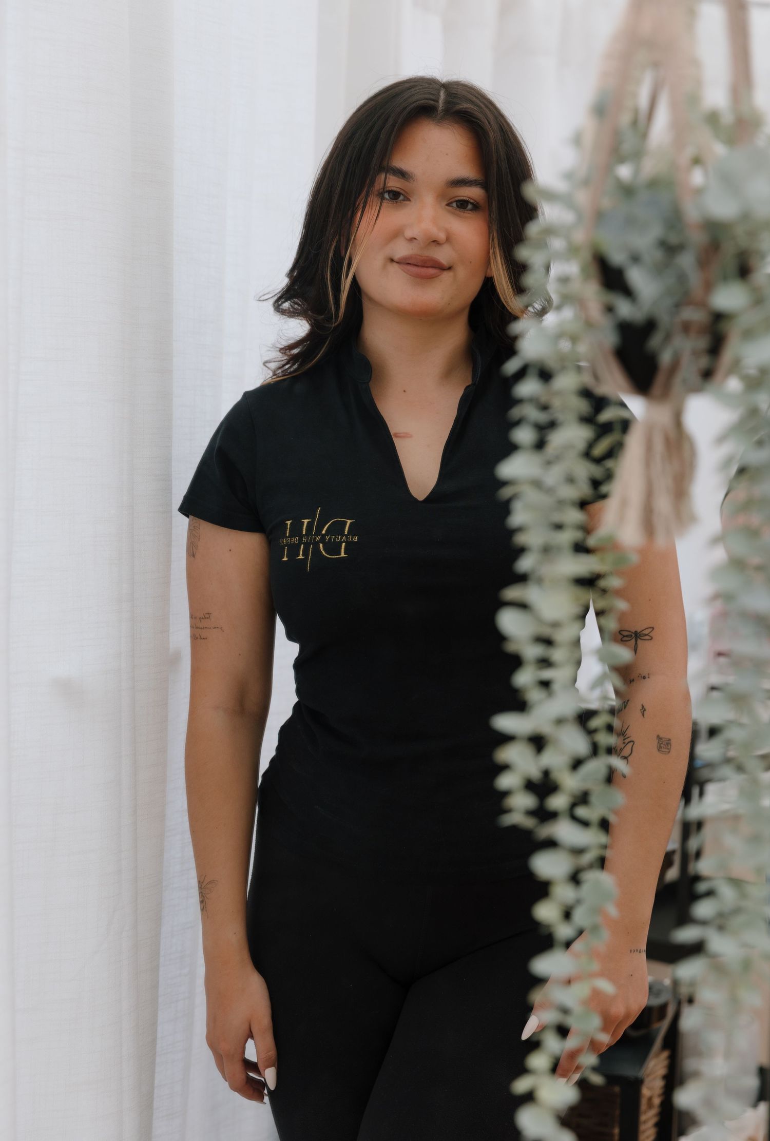 Person wearing all black outfit posing against white backdrop with greenery.