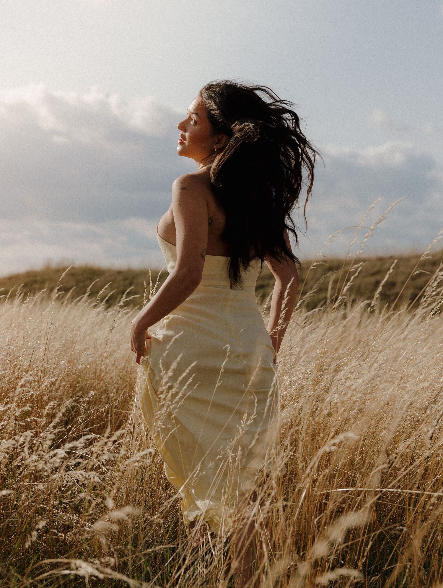 Woman in white dress standing in golden wheat field with wind-blown hair against cloudy sky.