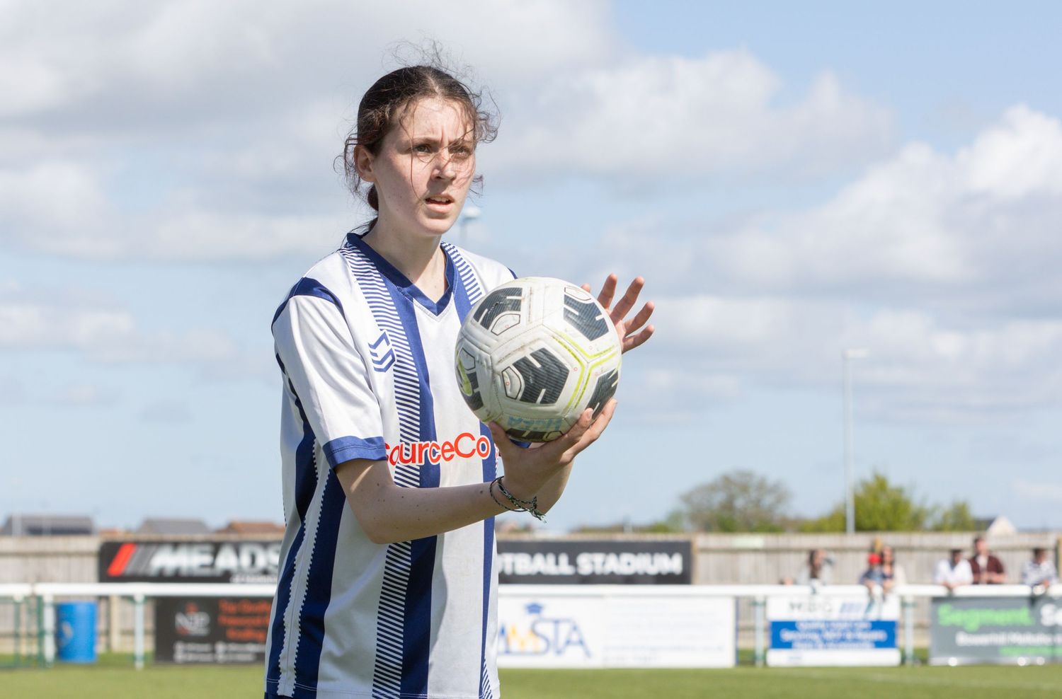 A soccer player in a blue and white striped jersey holds a football on a sunny day at a stadium.