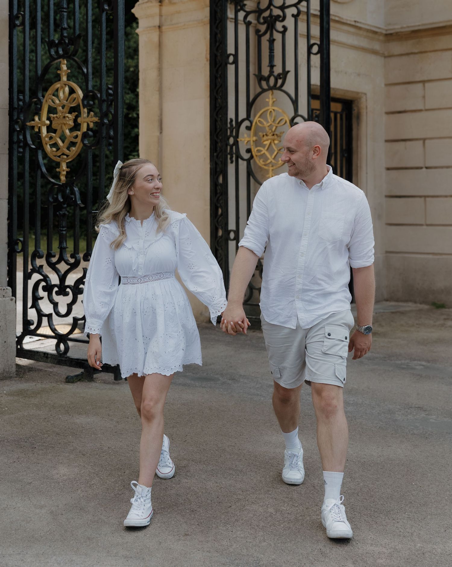 Couple in matching white outfits walking playfully near an ornate iron gate entrance.