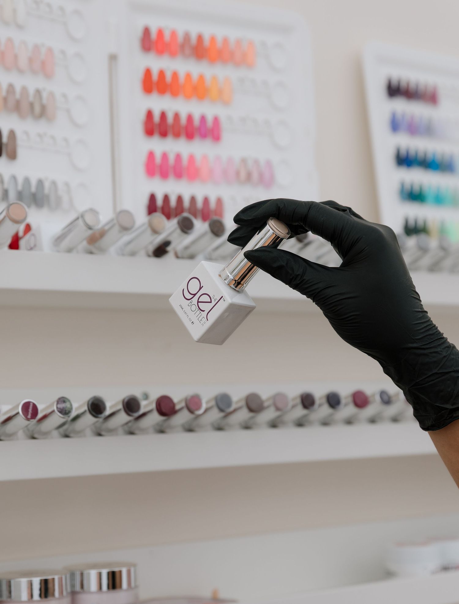Hand wearing black glove holding nail polish bottle in salon with colorful display wall.