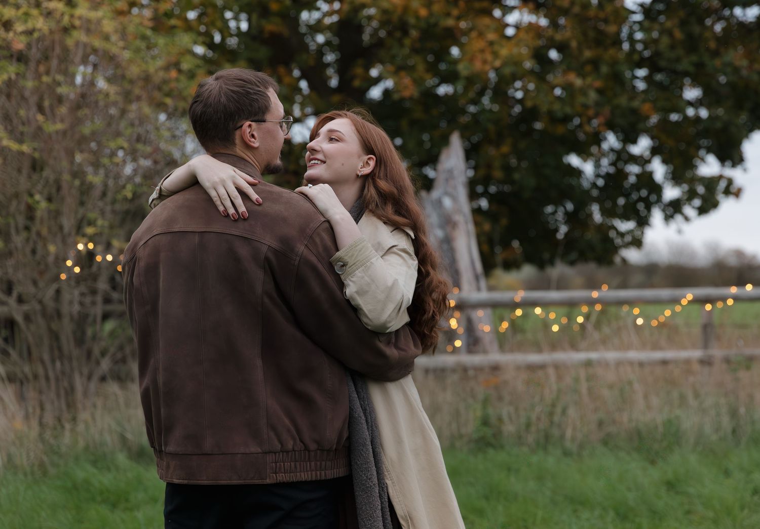 A romantic couple embraces and kisses in an outdoor setting with autumn trees and string lights in the background.