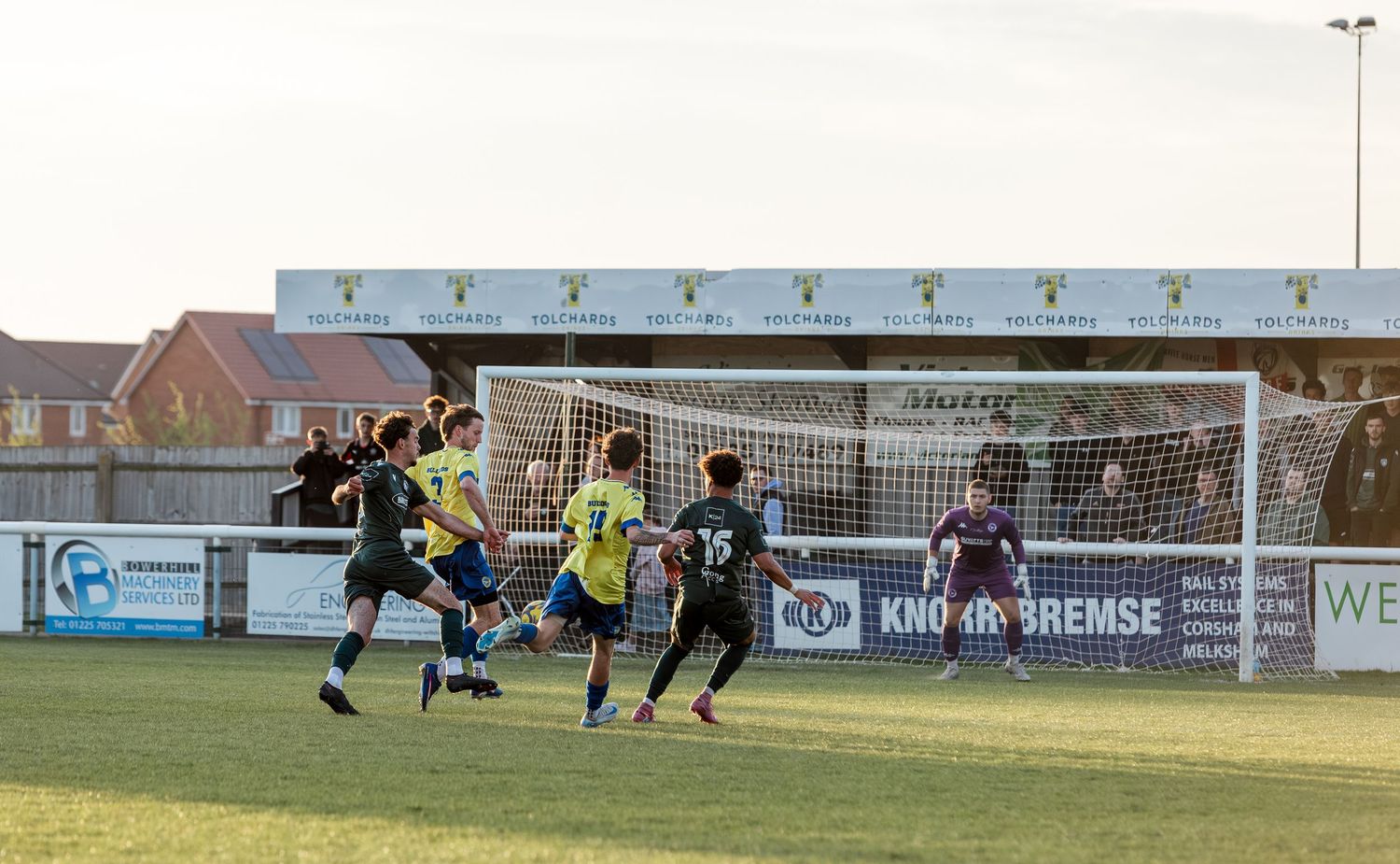 Soccer players compete near the goal during an outdoor match at a local football ground on a sunny day.