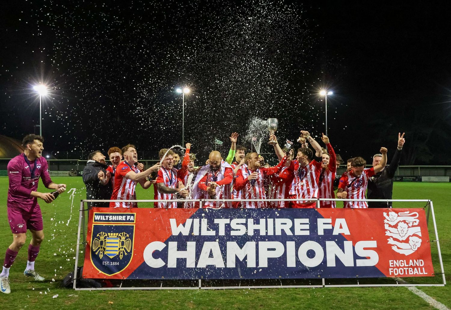 Soccer team celebrates with trophy as Wiltshire FA Champions under stadium lights.