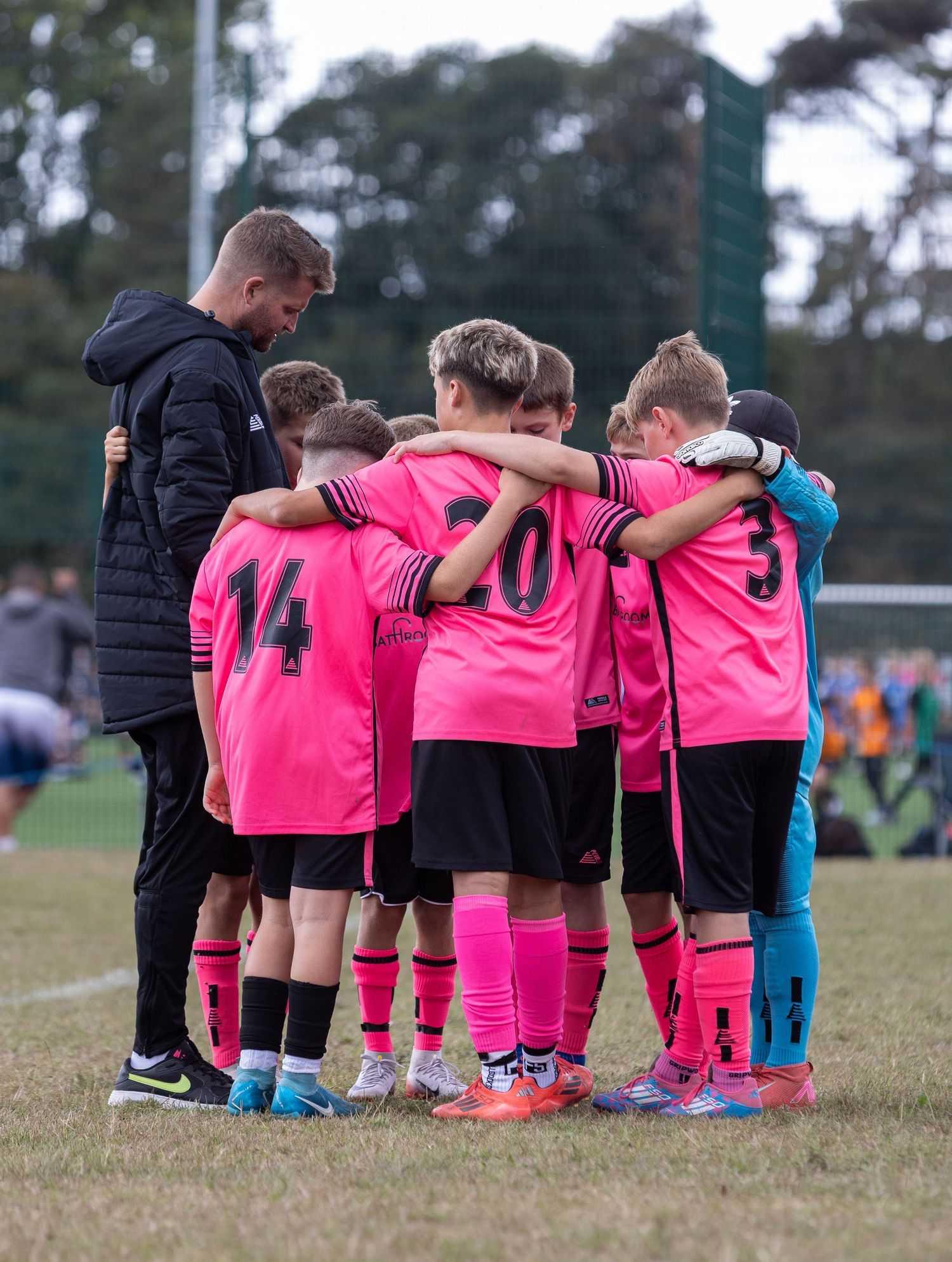 Youth soccer team huddles together in bright pink jerseys during a match on a grass field.