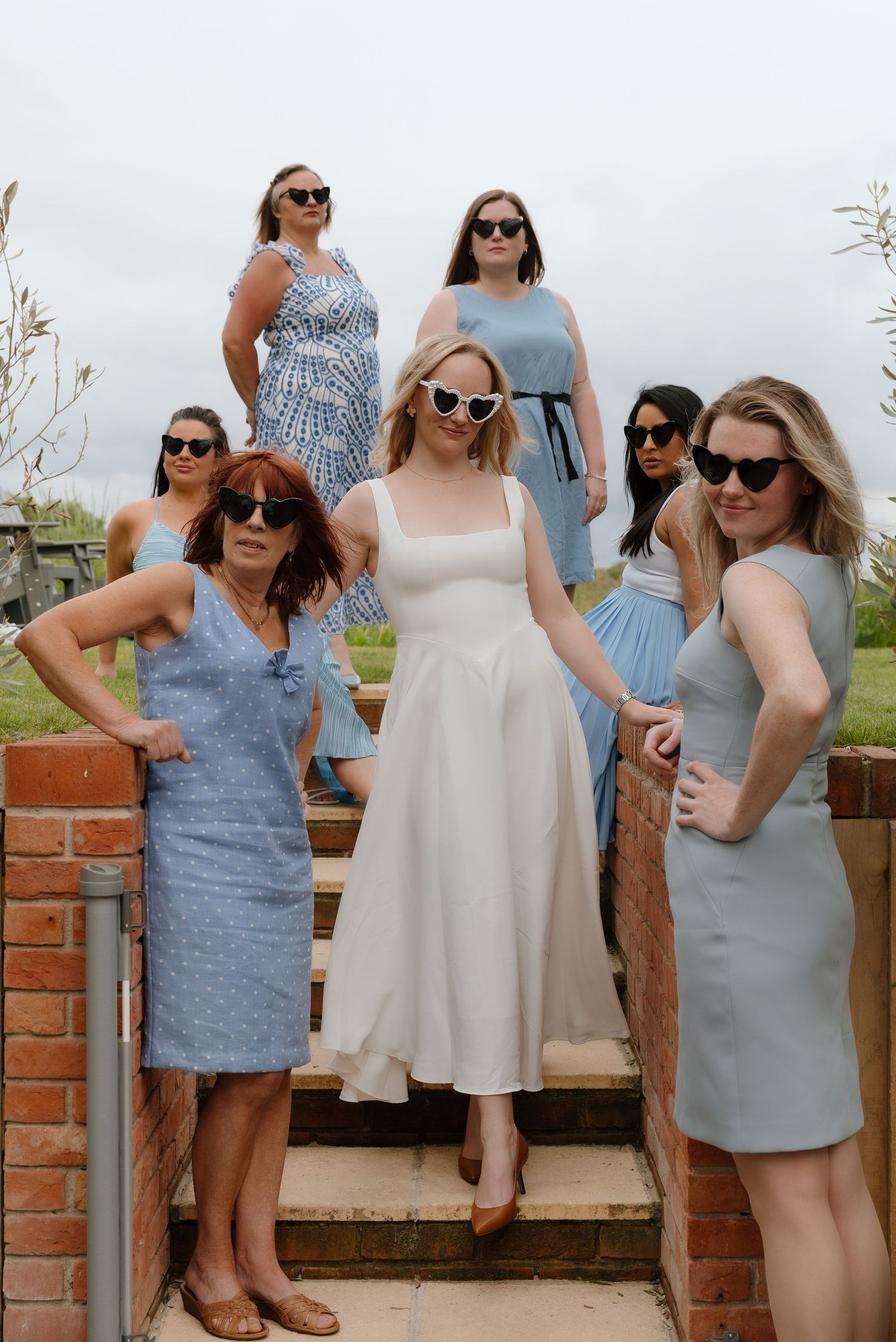 Group wearing sunglasses and summer dresses pose casually against a brick wall outdoors.