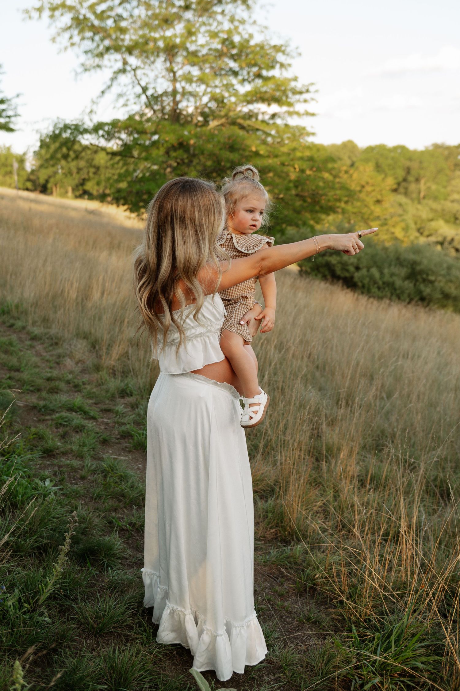 A mother in a white maxi dress holds her toddler girl in a sunlit field, as the child points into the distance.