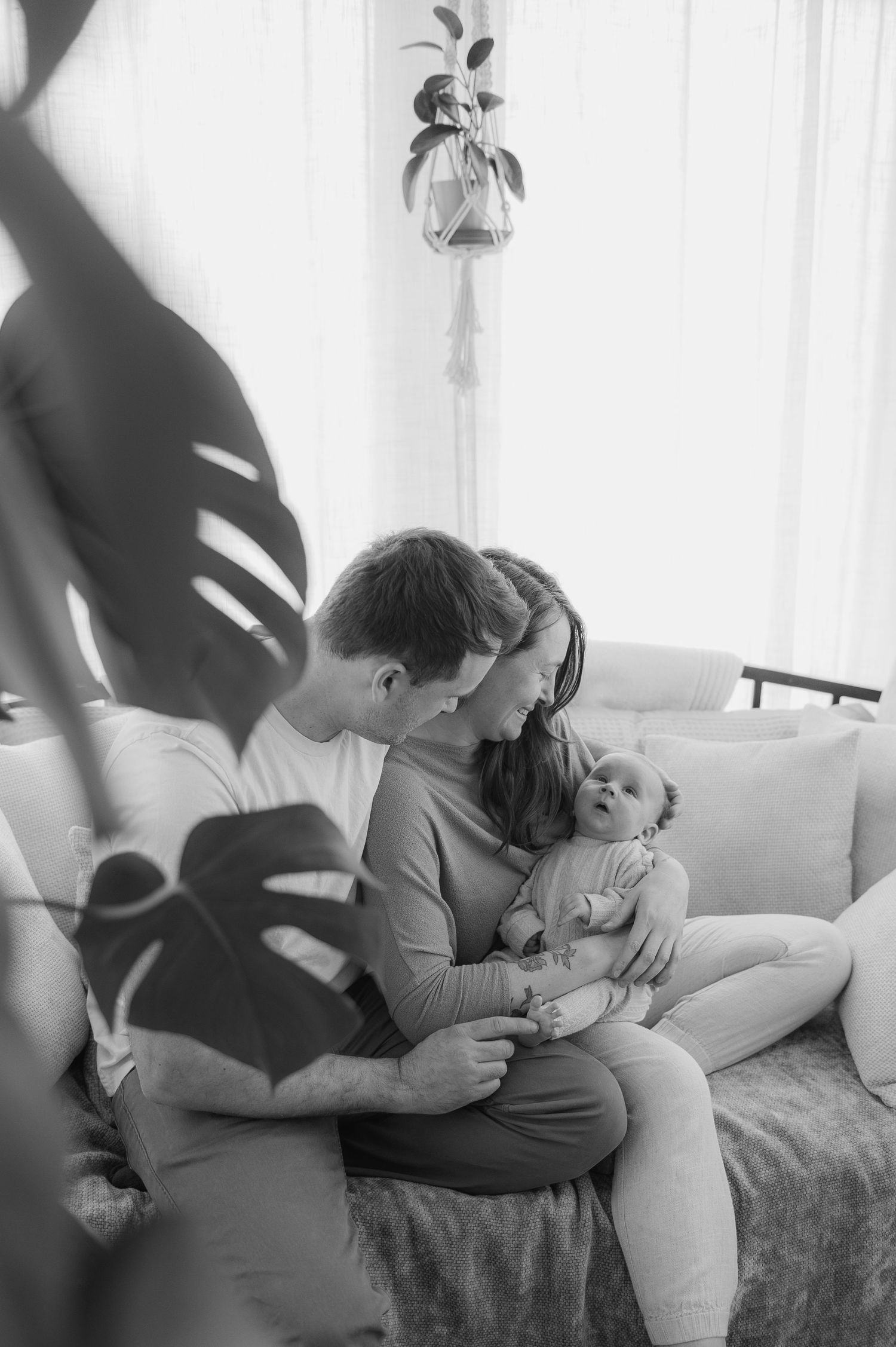 Black and white photo of parents sitting on a couch, lovingly gazing at their newborn baby.
