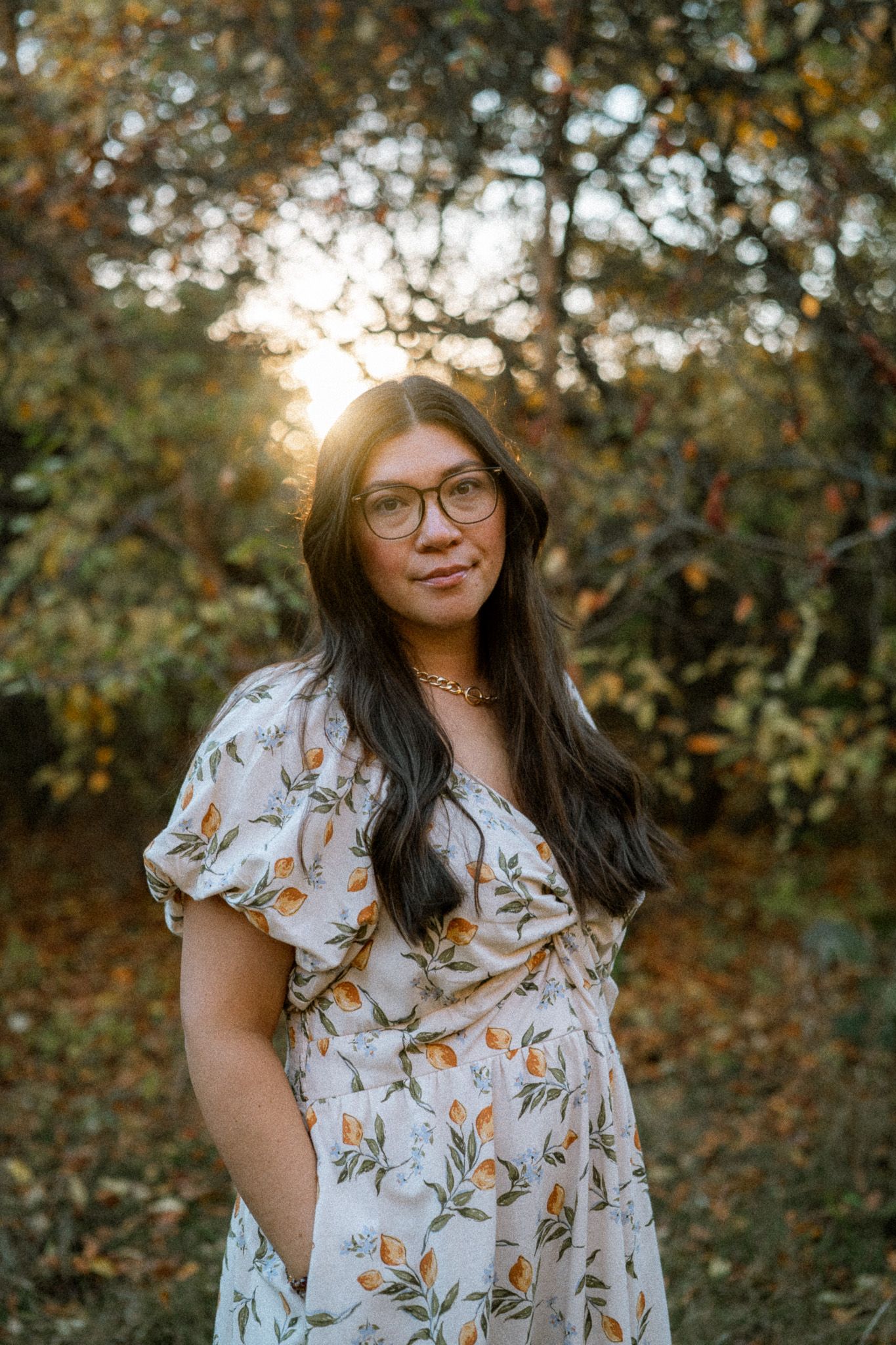 A person wearing glasses and a floral dress stands in a sunlit autumn forest setting with golden bokeh lighting.