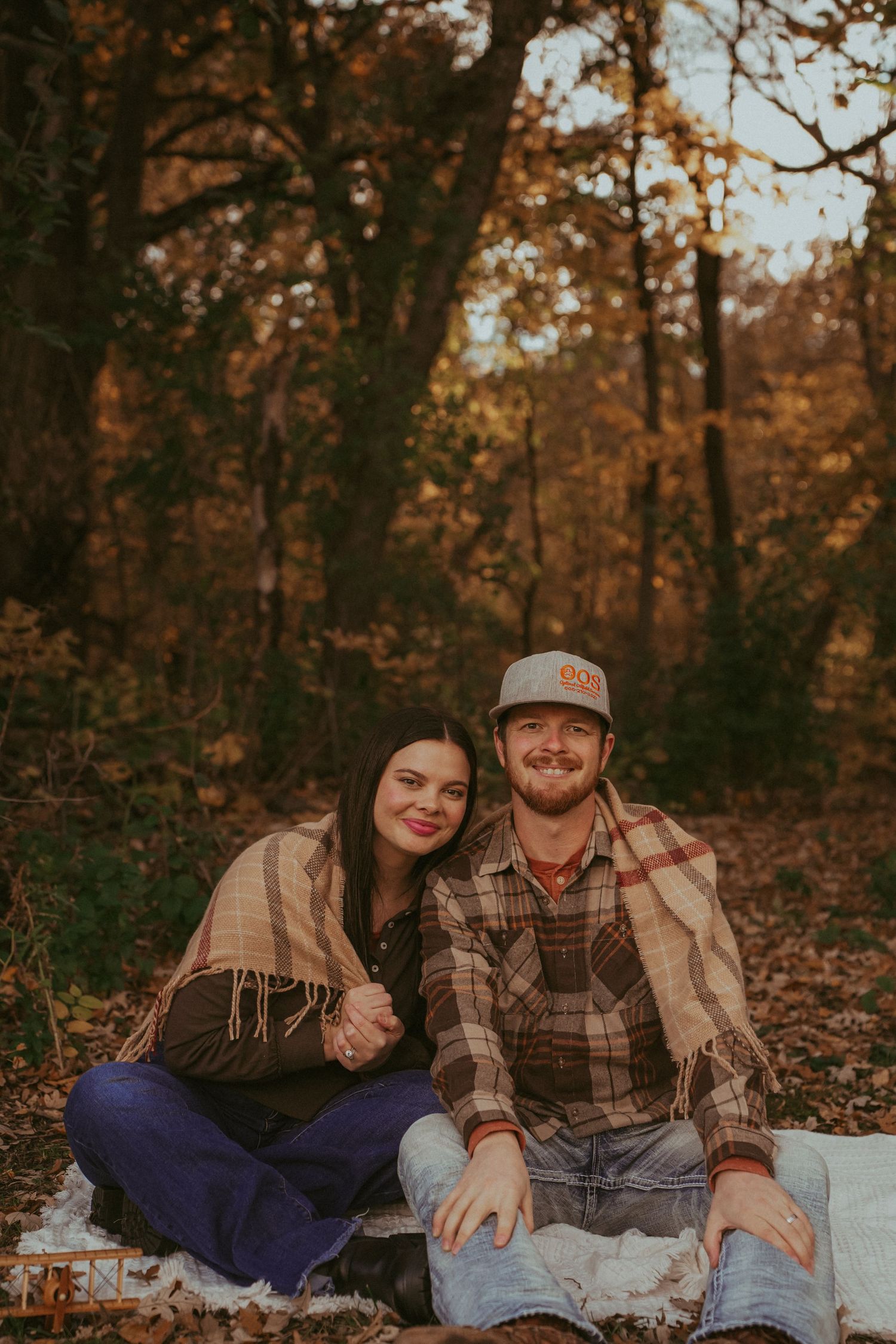 A couple sits on a blanket in autumn woods during golden hour wearing casual plaid and fringe clothing.