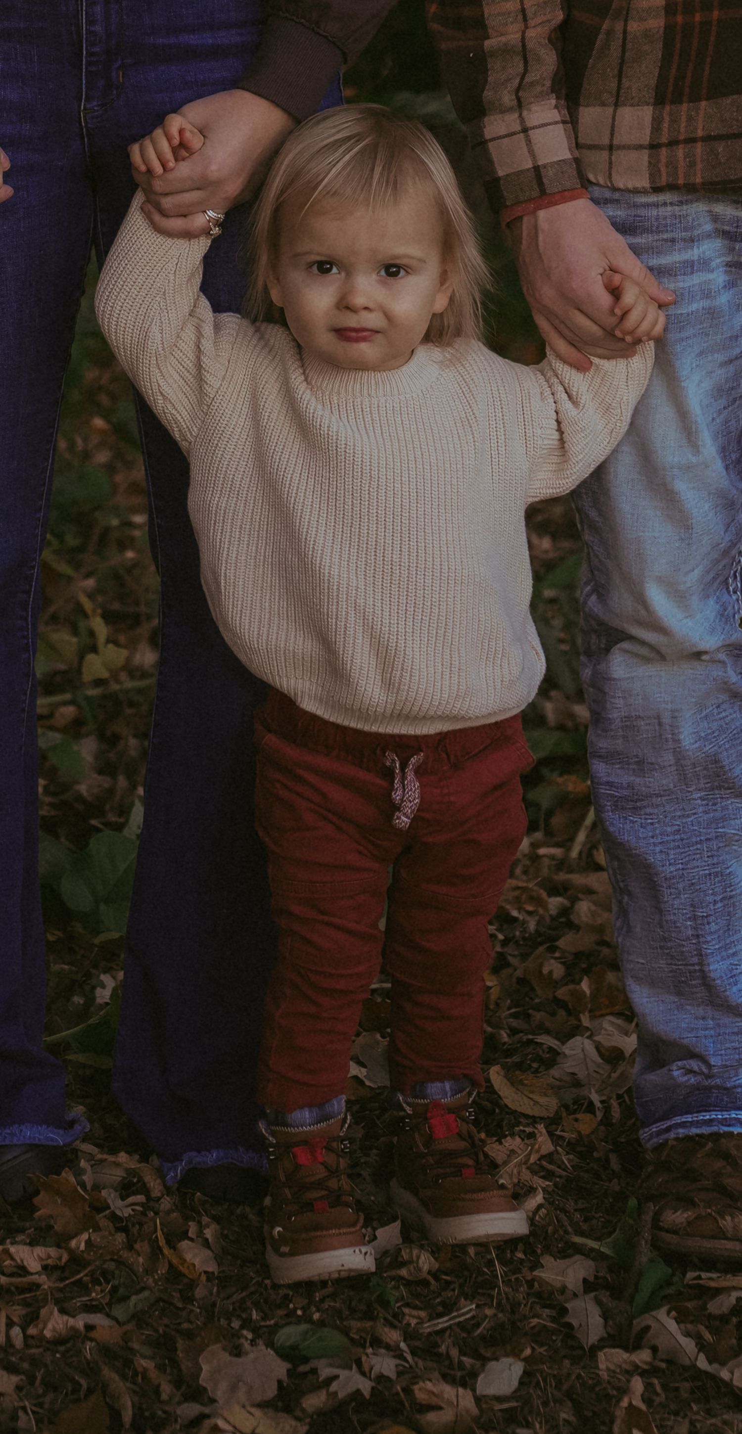Small child in cream sweater and burgundy pants stands against dark background.