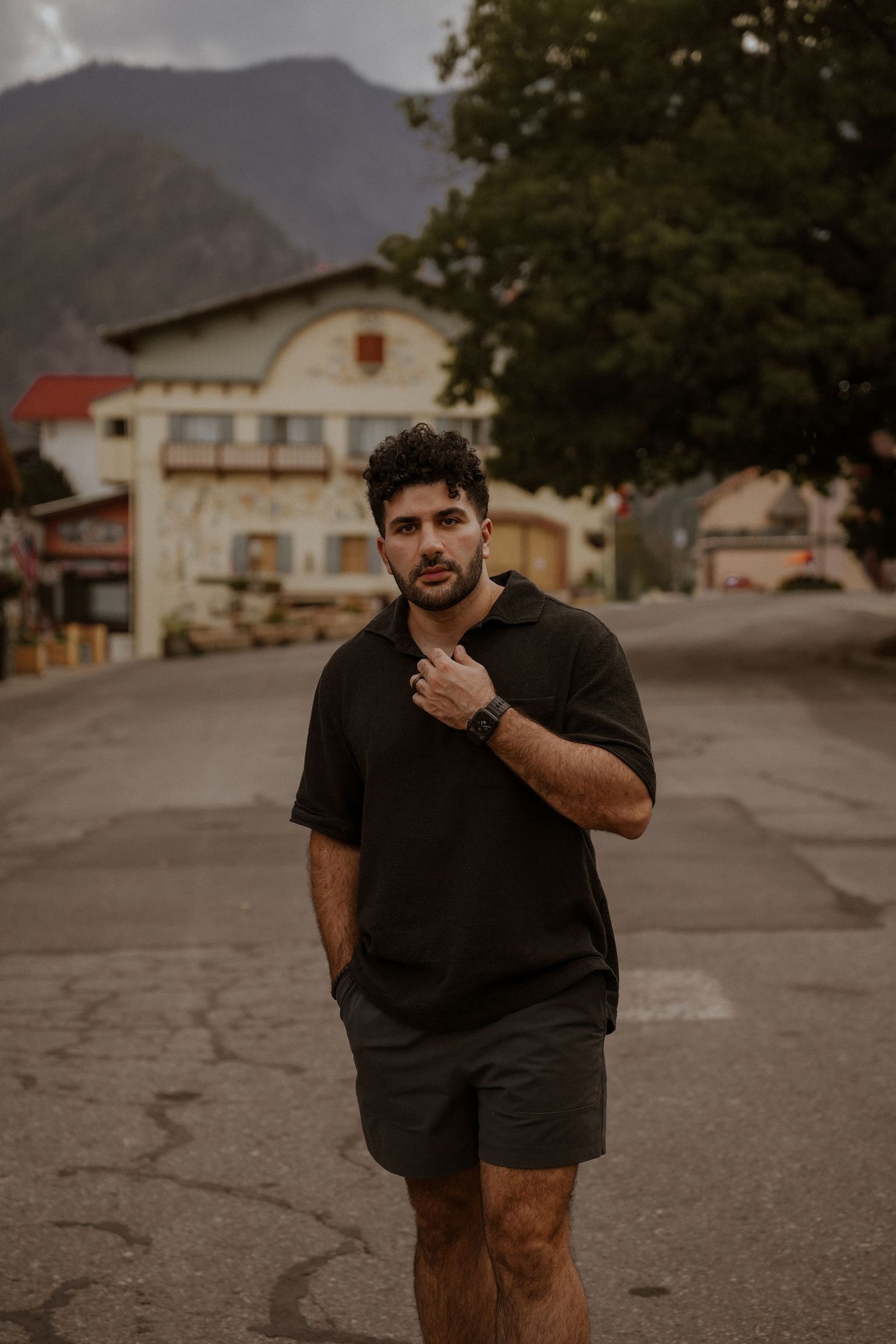 Person in black clothing poses thoughtfully on an empty street with mountain backdrop.
