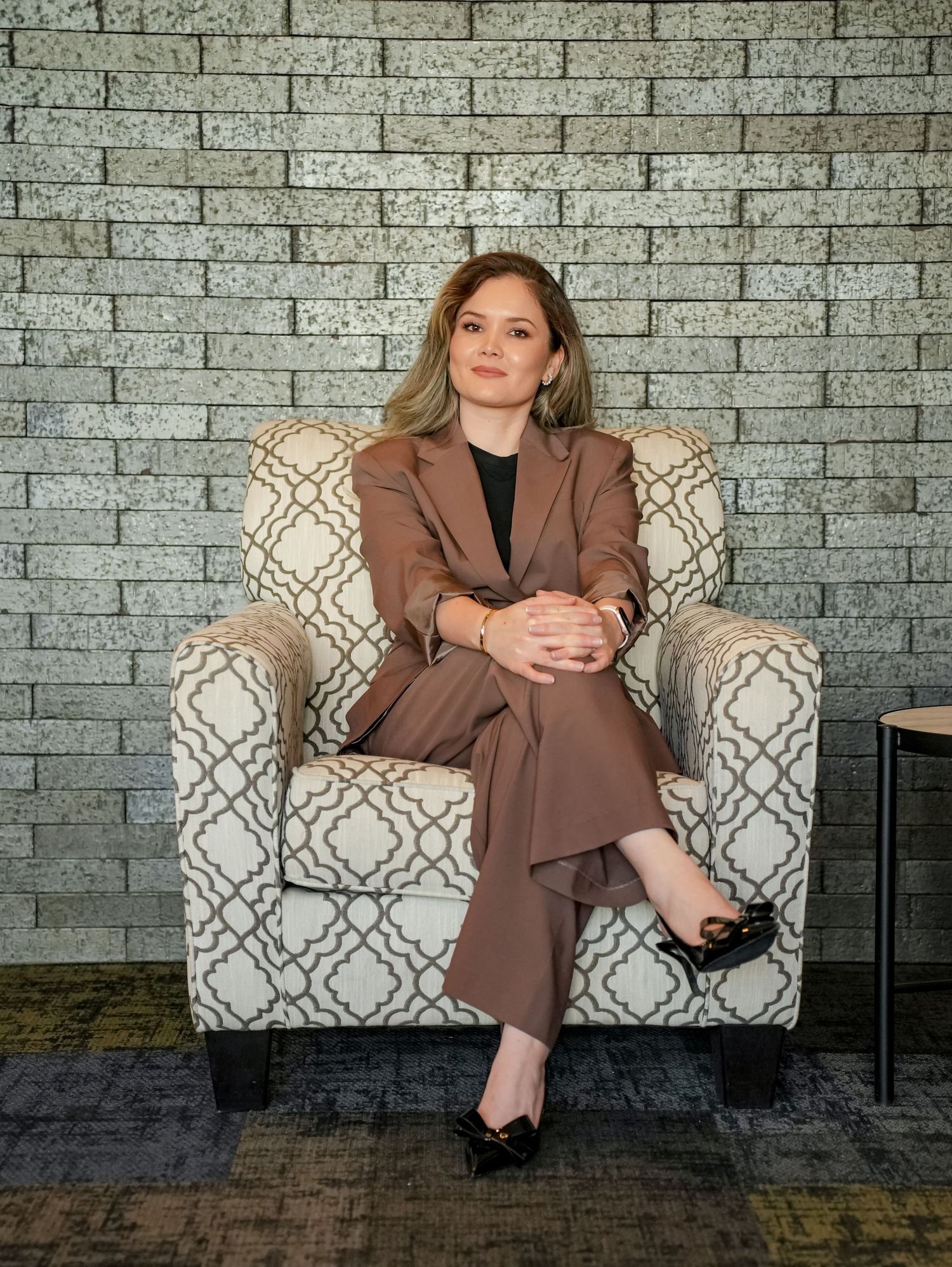 Professional seated in a brown suit on a patterned accent chair against a gray brick wall.