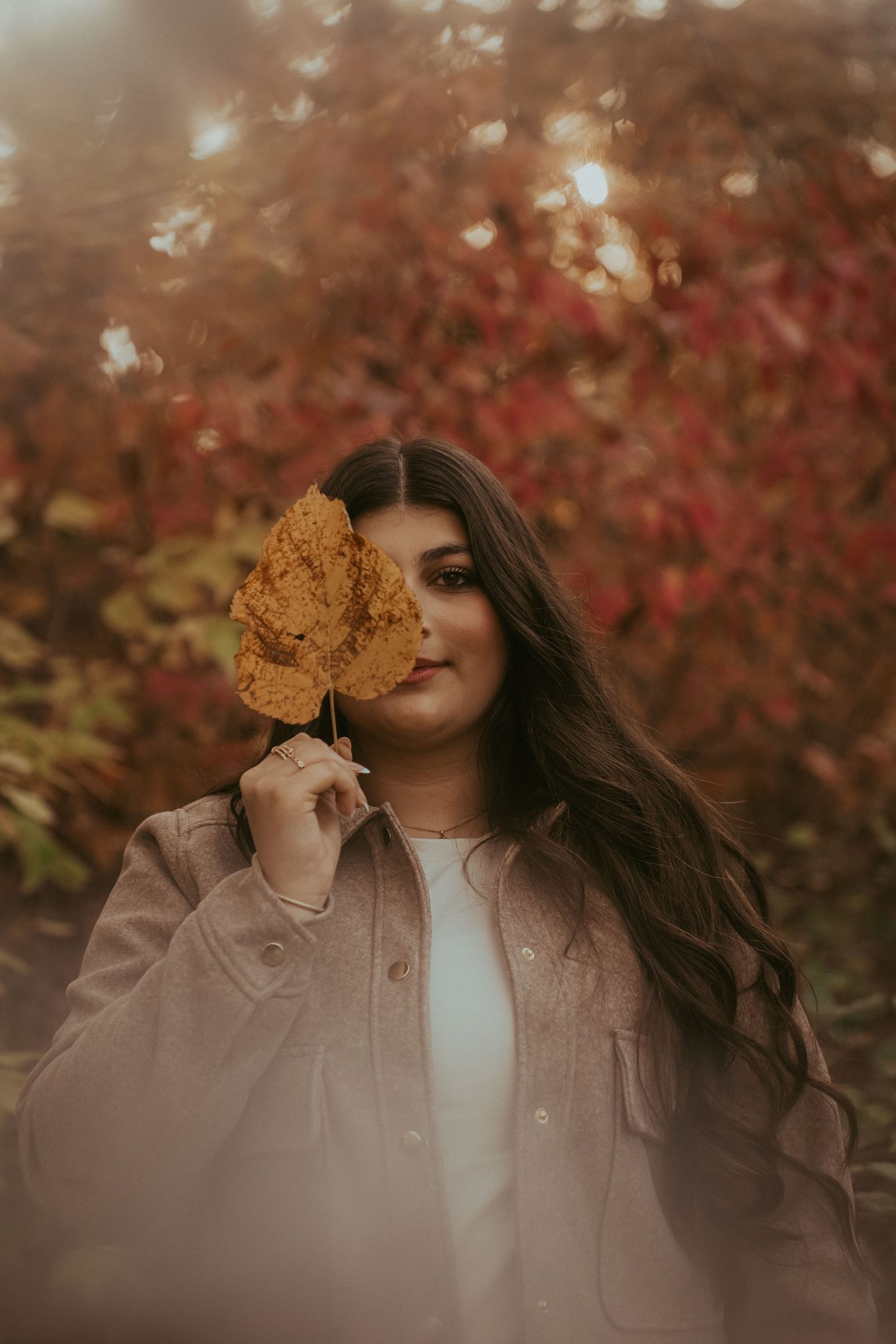 A person in a beige jacket holds up a golden autumn leaf against a backdrop of red fall foliage.