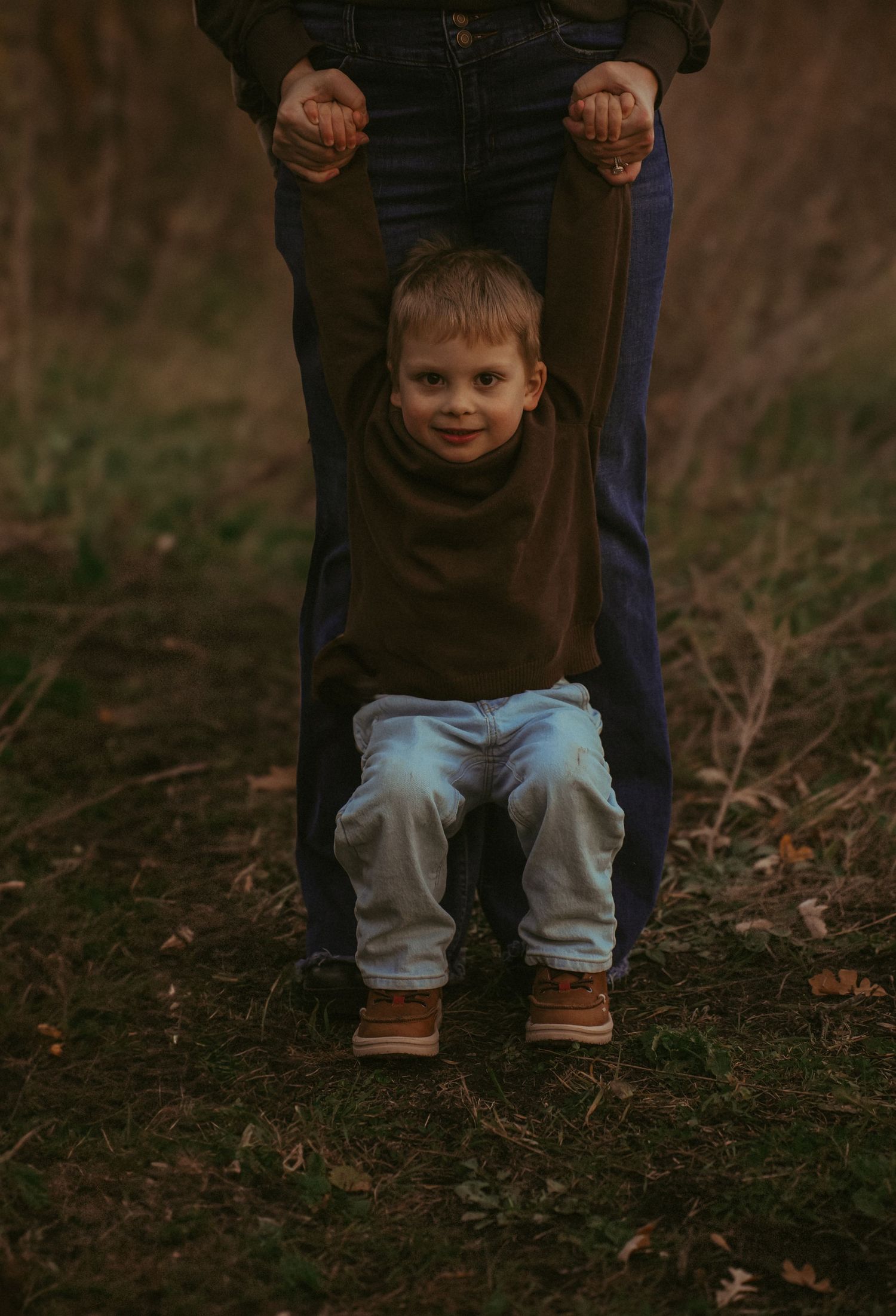 Child swings joyfully while being held by adult hands against natural background.
