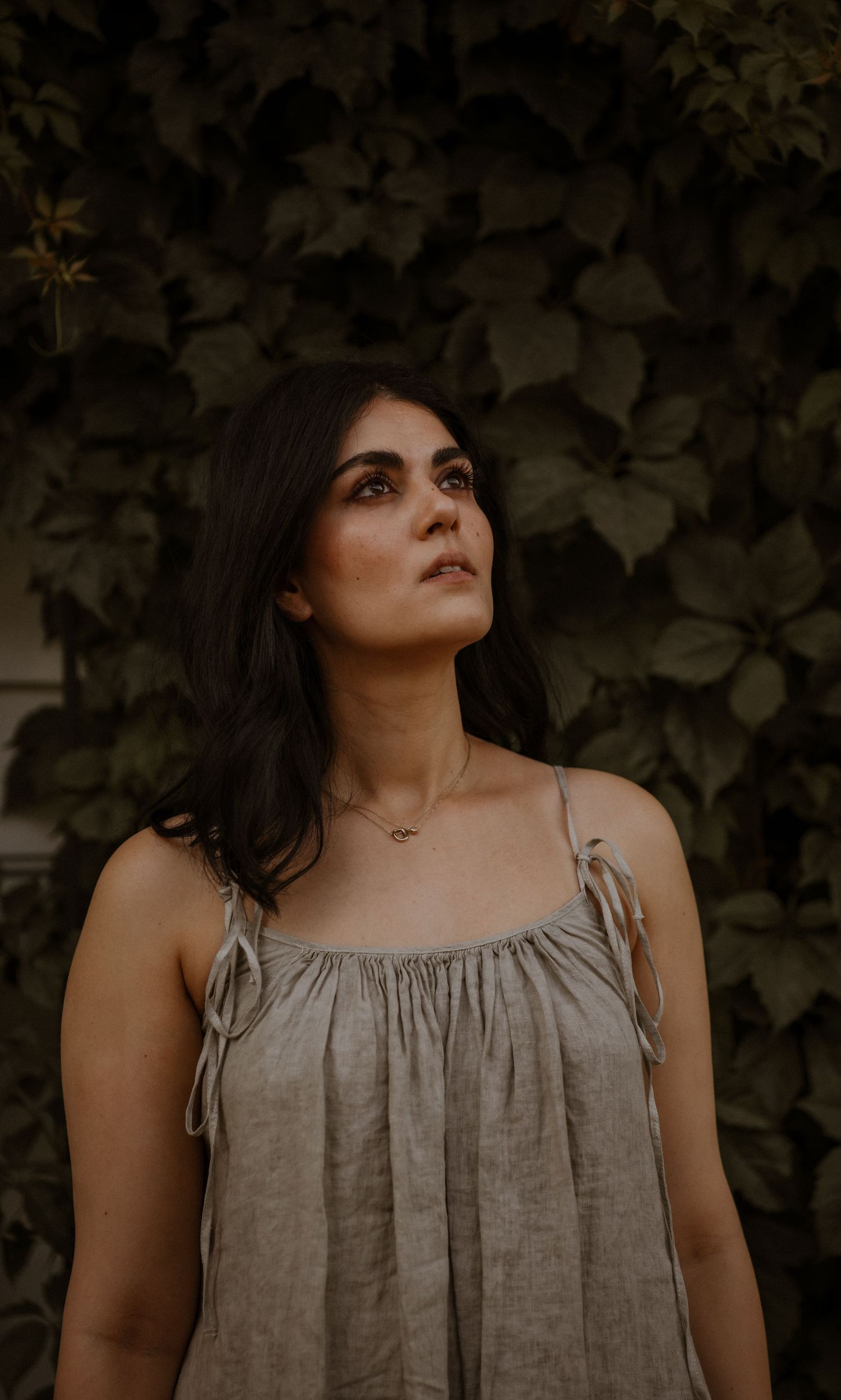 Person wearing a gray linen dress poses against a leafy backdrop in warm evening light.