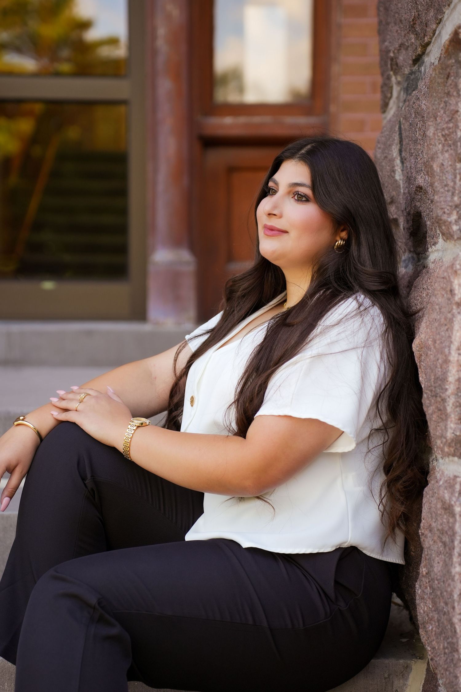 Woman in white blouse and black pants sits casually against stone wall outdoors.
