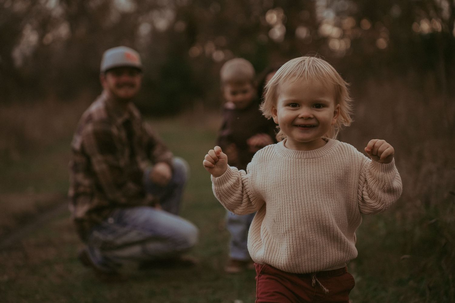 A toddler in a cream sweater smiles while playing outdoors at sunset.