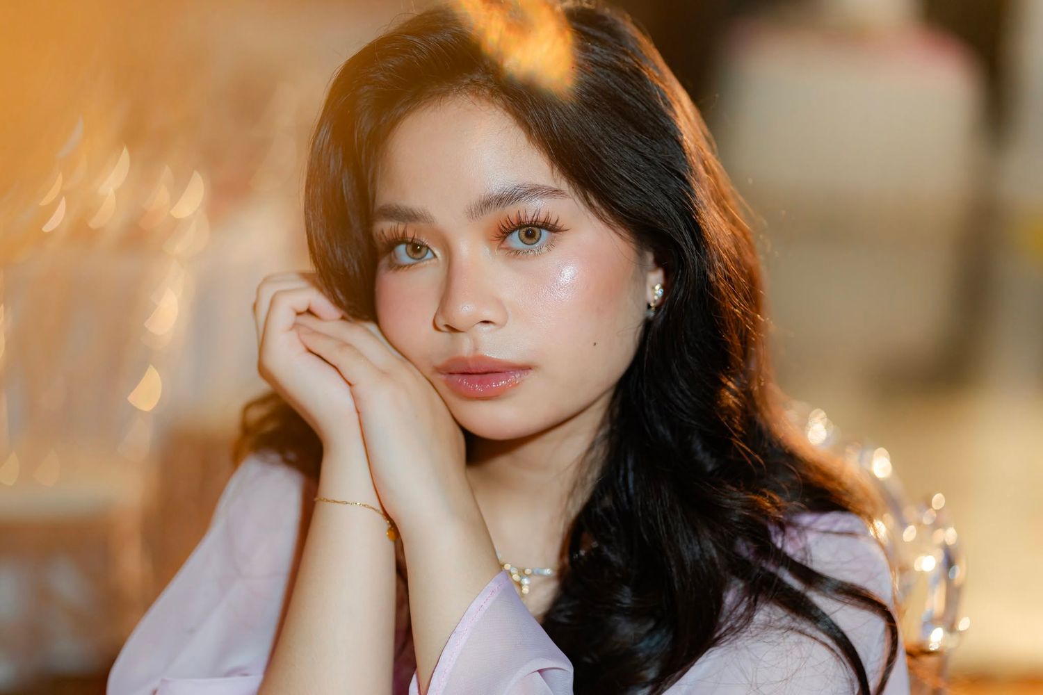 Young woman with wavy dark hair and natural makeup poses gracefully with hands clasped near her face.