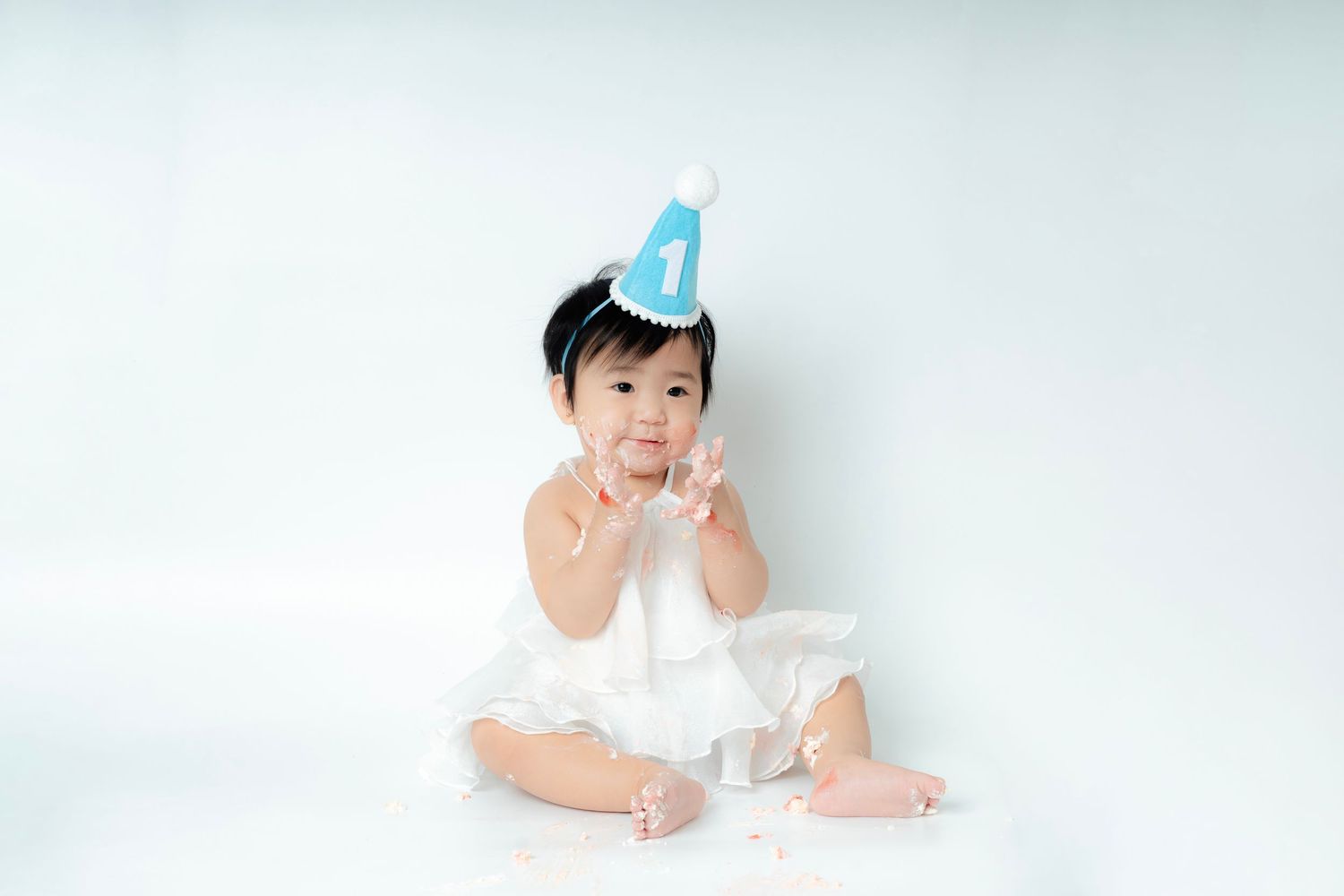 Baby wearing blue party hat and white dress sits on floor during first birthday photo session against white backdrop.