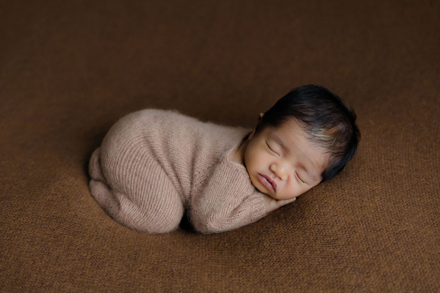 Newborn sleeping peacefully wrapped in knit fabric on brown textured background.