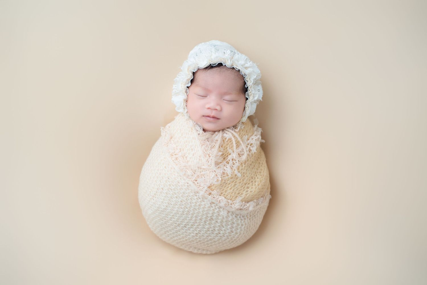 Sleeping newborn in cream knit wrap with white lace bonnet against neutral backdrop.