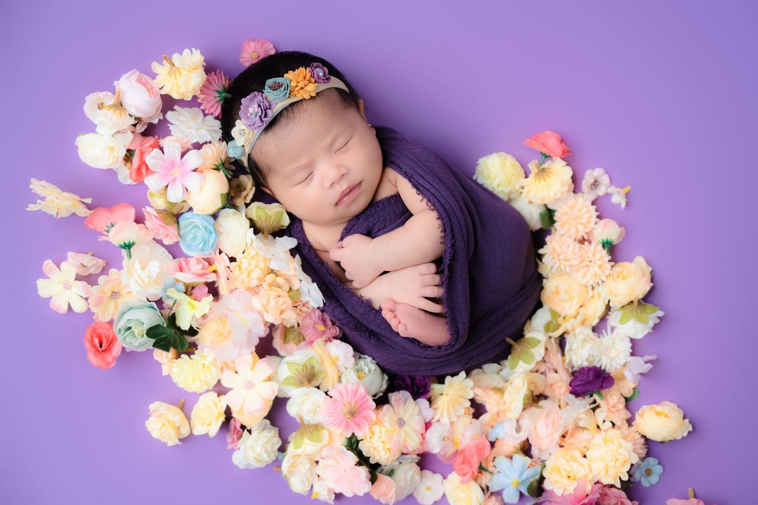 Newborn wrapped in purple fabric surrounded by colorful spring flowers on purple background.
