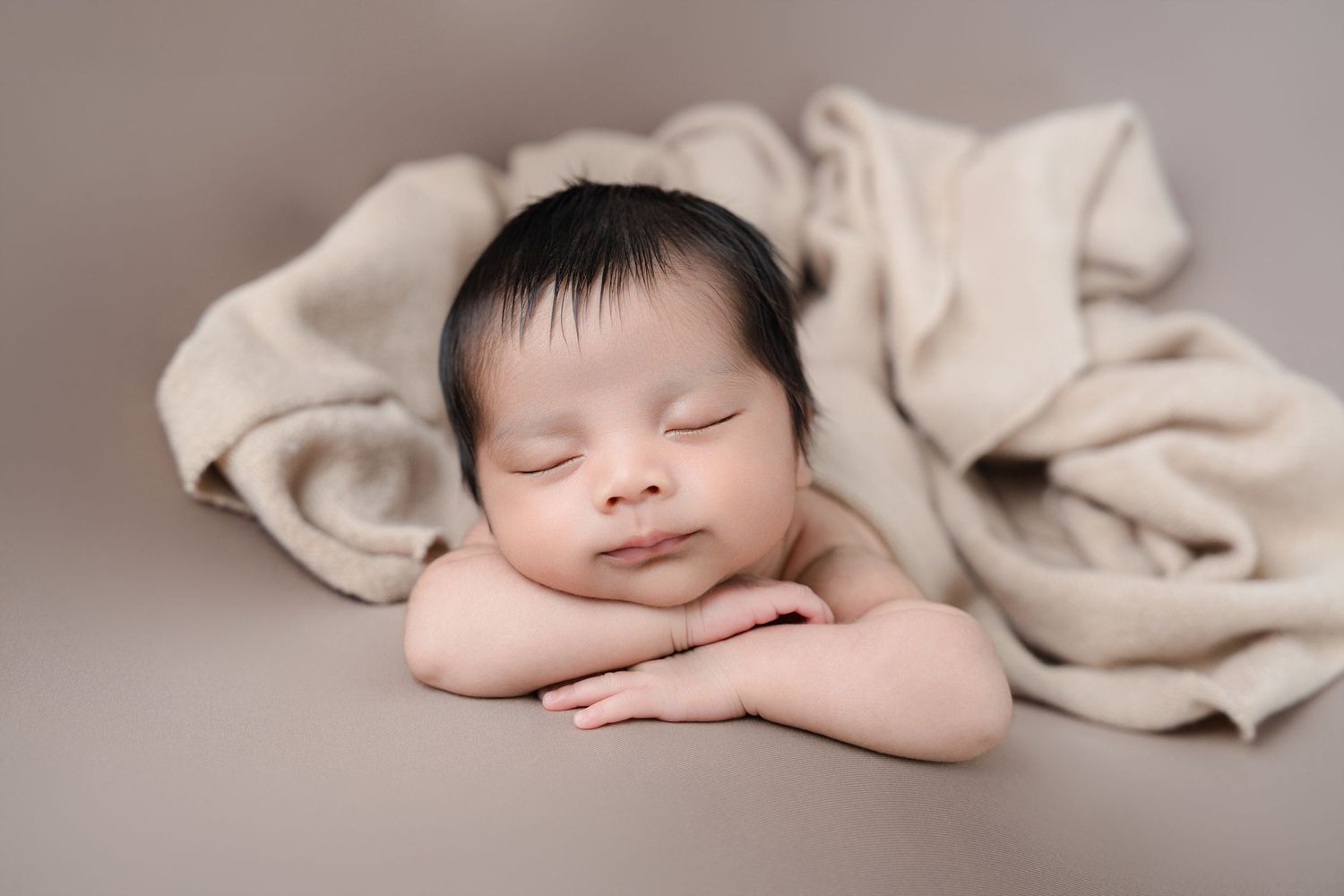 Peaceful sleeping newborn resting head on folded arms with tan blanket.