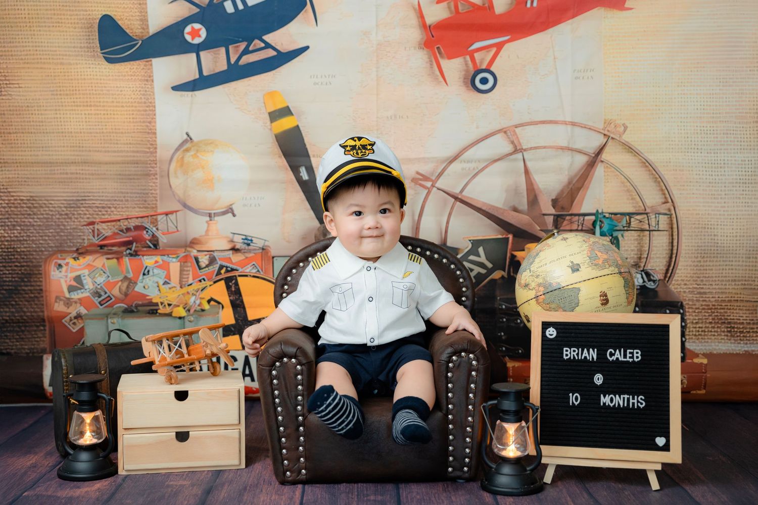 Baby photo session with vintage aviation theme featuring toy planes and wooden props on beige background.