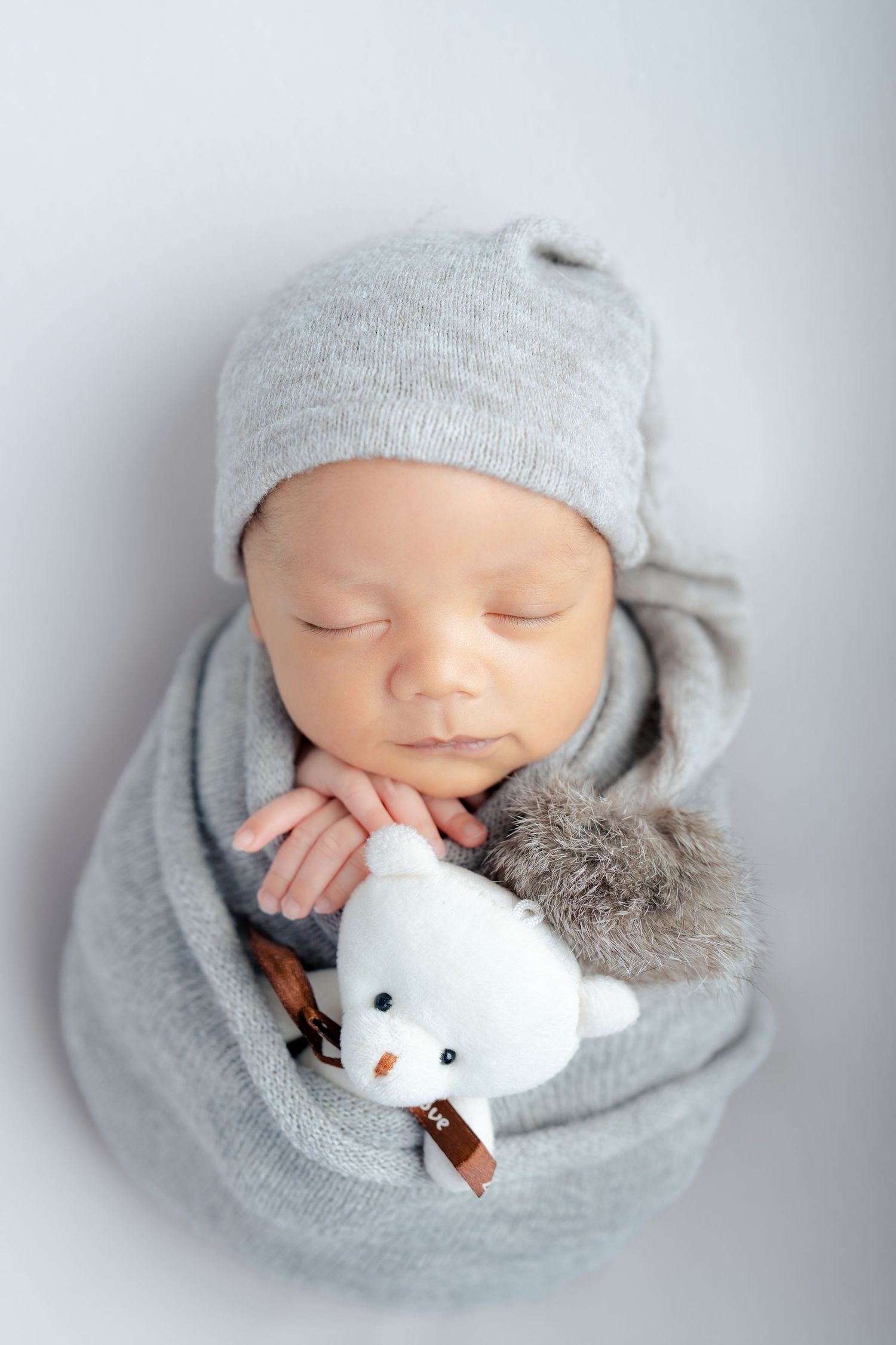 Sleeping newborn wrapped in soft gray swaddle with knit hat and white plush toy bear on white background.