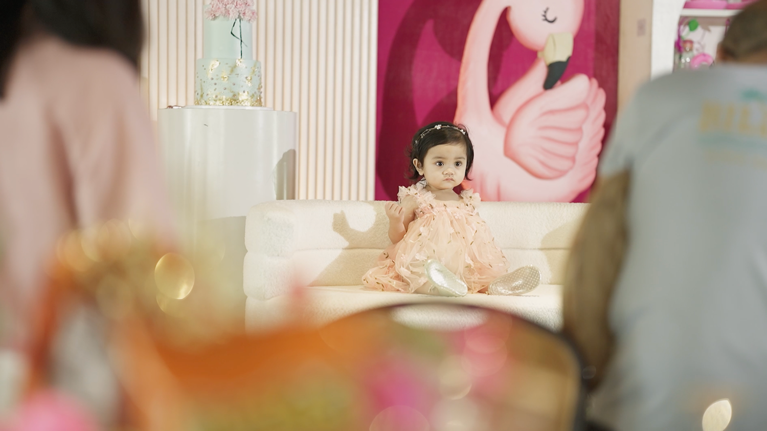 A young toddler girl in a pink dress sits on a white sofa at a birthday party with flamingo decor.